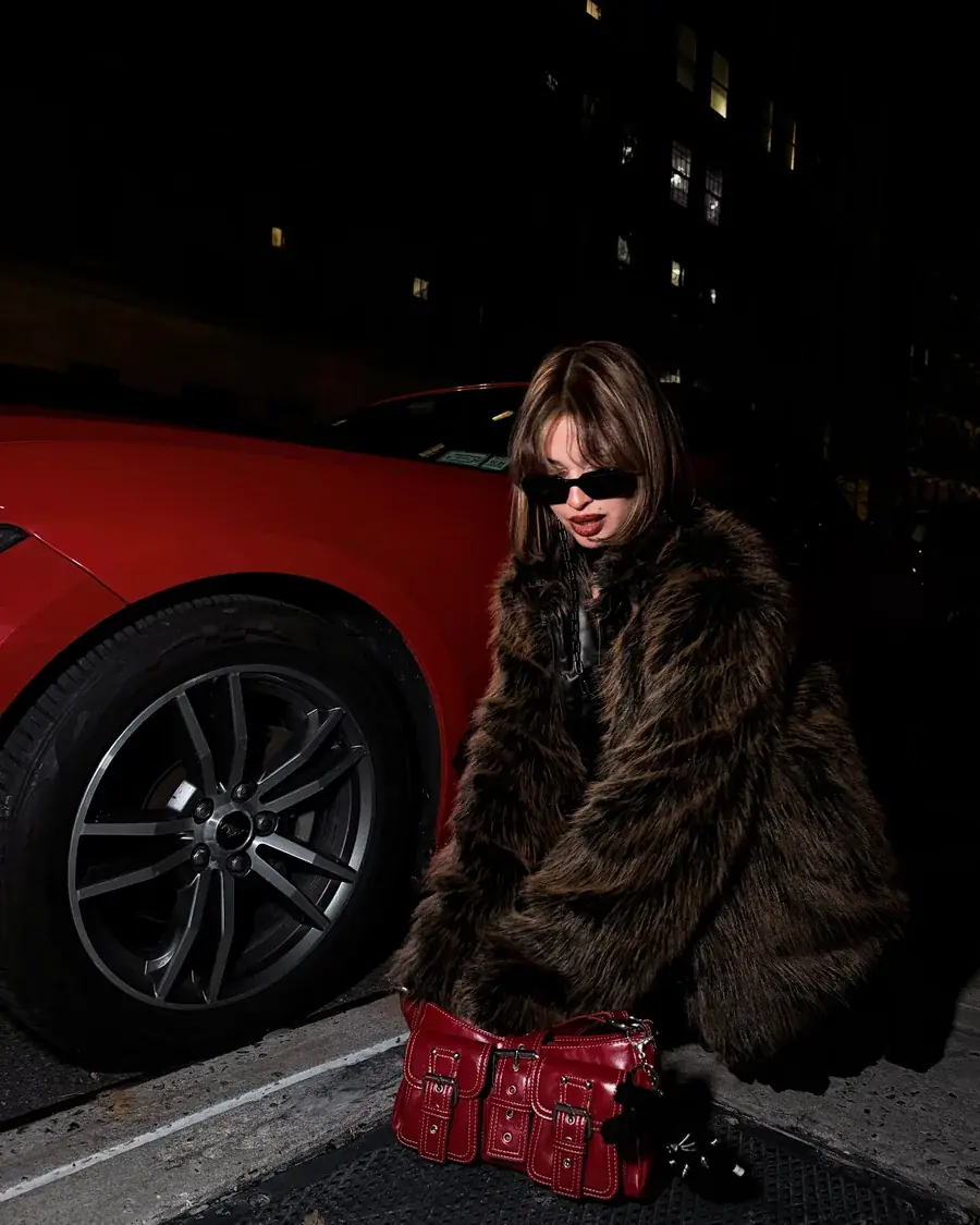 Modelo con gafas de sol y abrigo de piel agachada junto a un Mustang rojo, de noche, con un bolso rojo de charol